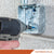 Person using a drill to tighten a screw into a metal box on a concrete wall, with 'Fasteners Plus' branding.